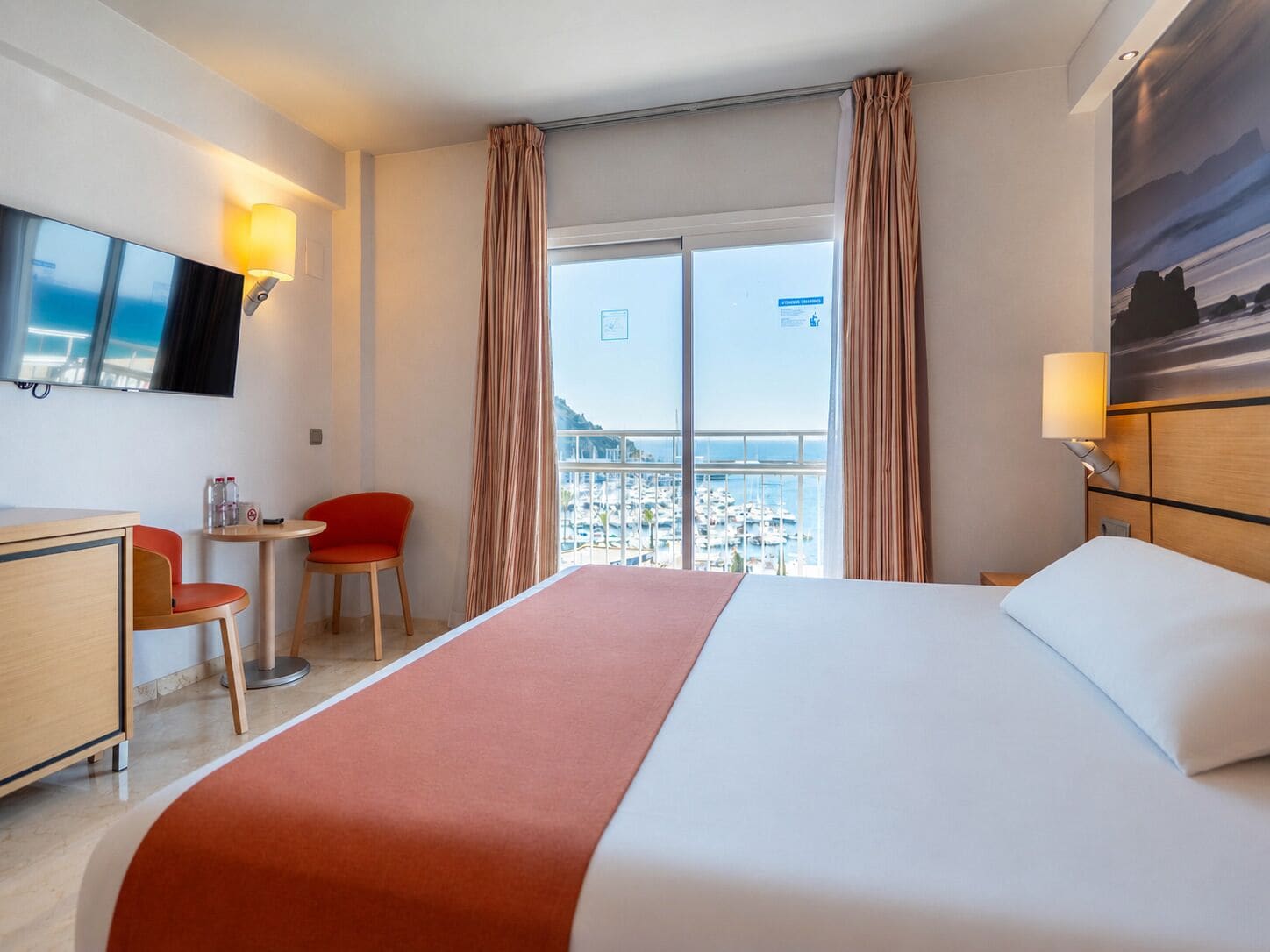 A hotel room with a bed, a desk, a chair, a TV, and a view of the marina.