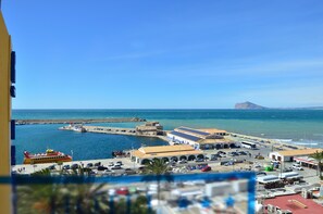 Un pueblo costero con un puerto deportivo, una montaña imponente al fondo y diversos edificios, incluyendo una estación de autobuses y comercios.
