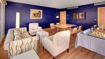 Una habitación con pared azul, suelo de madera, sofá blanco, sillón blanco, una mesita, una mesa de comedor más grande con sillas, un televisor y un aparador con comida.