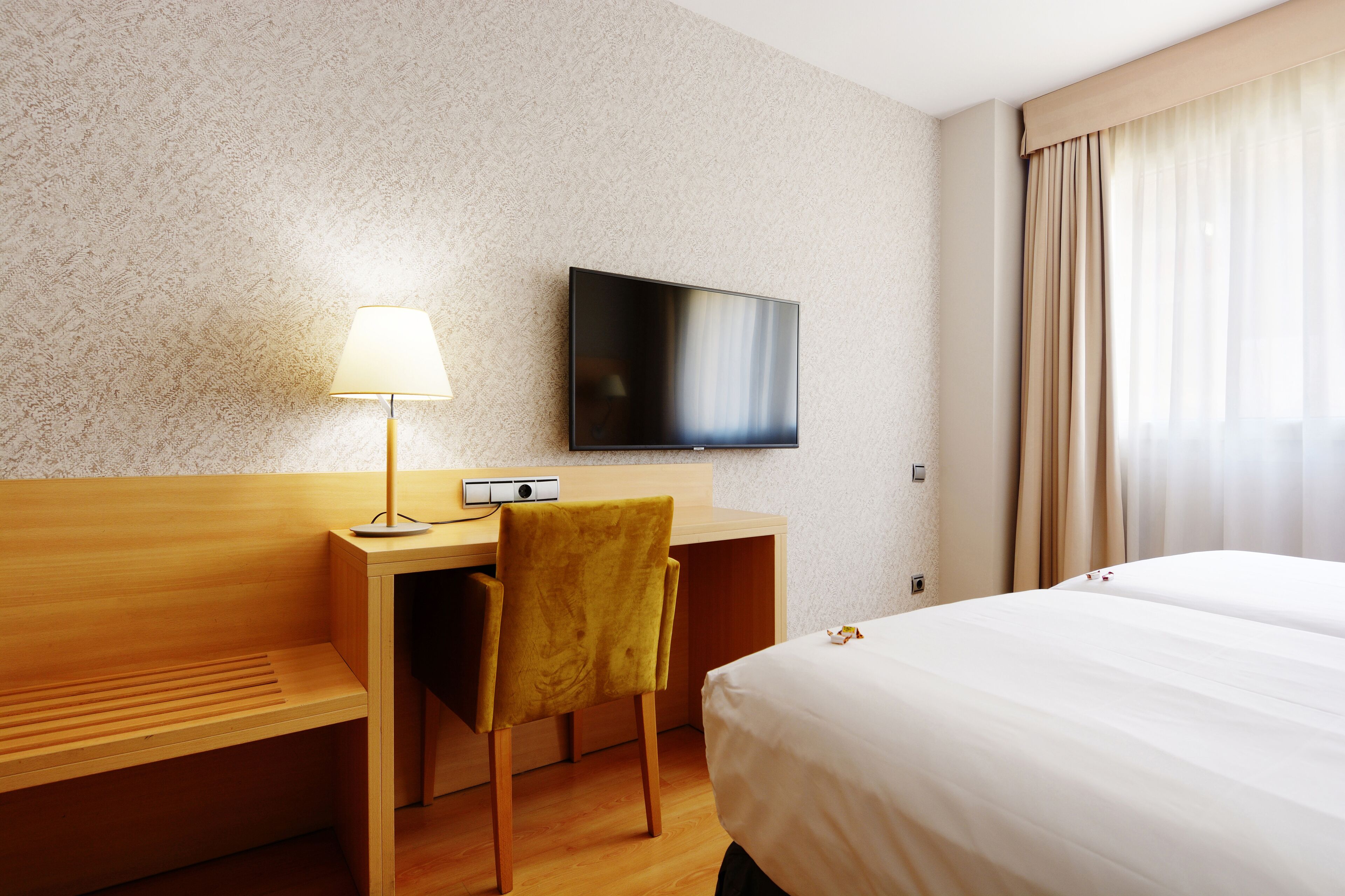 A hotel room with a bed, a desk with a lamp, a television, and a window with curtains.