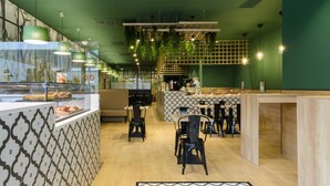 Interior de restaurante con pared verde, salpicadero de azulejos estampados, plantas colgantes y mesas y sillas de madera.