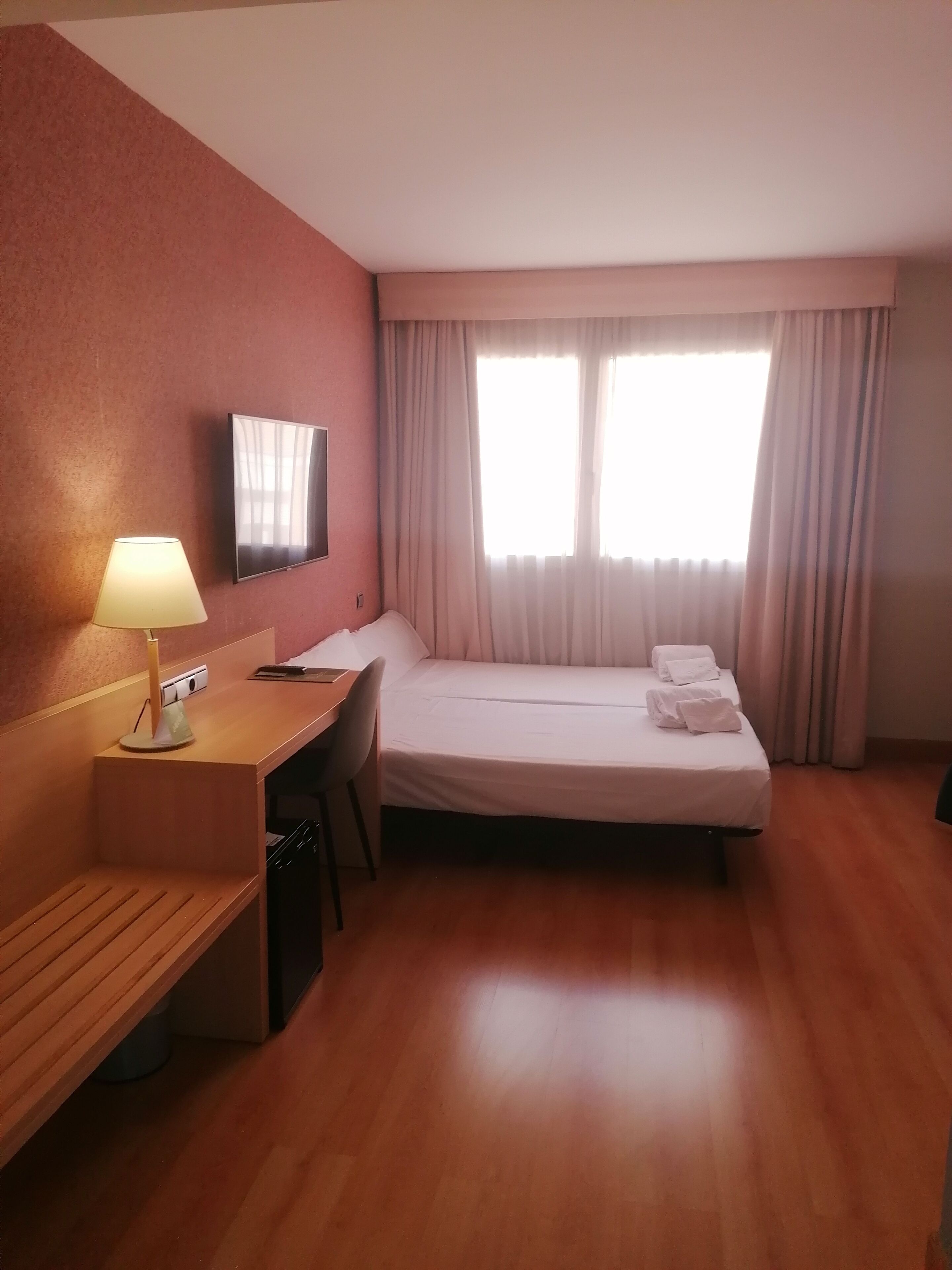A hotel room with a bed, a desk, a chair, a TV mounted on the wall, and a window with curtains.
