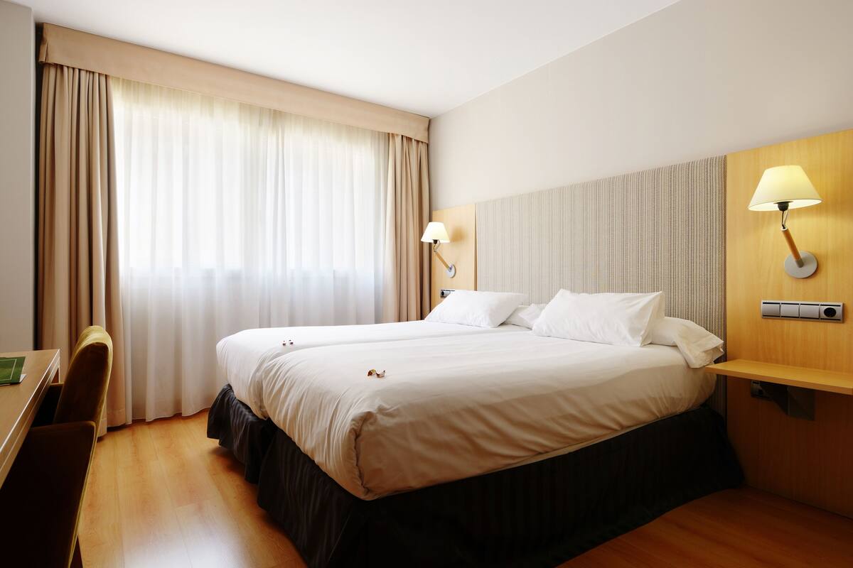 Habitación de hotel con dos camas, un escritorio, una silla, una lámpara y una ventana con cortinas.