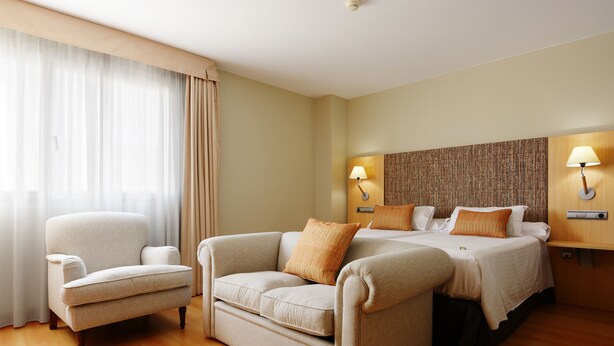 Habitación de hotel con sofá, cama y sillón. Dos lámparas de pared y un panel decorativo en la pared sobre la cama.