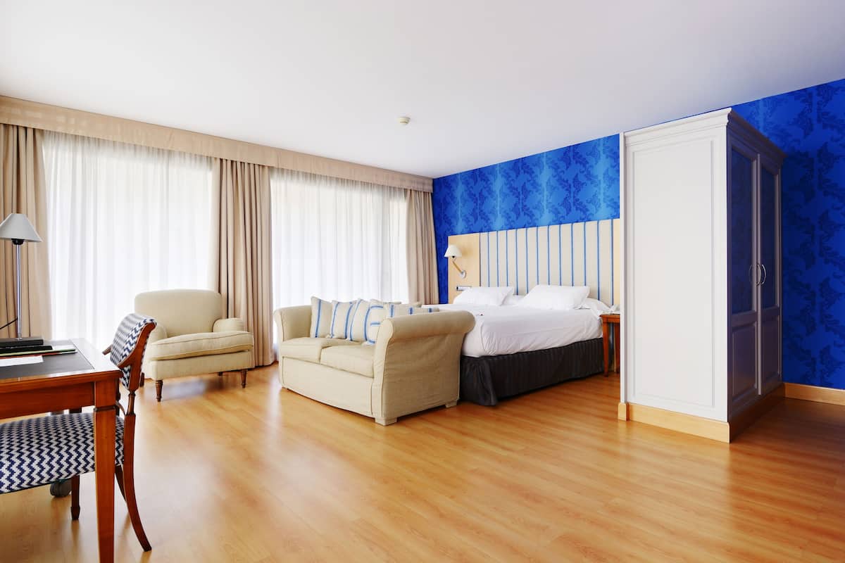 Una habitación de hotel con piso de madera, una cama con ropa blanca, un sofá color crema, un escritorio con lámpara y un armario.