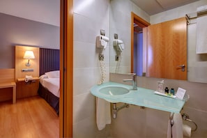 A hotel room with a bed, bedside table, lamp, and a bathroom with a glass sink, mirror, and towel rack. - U Hotel Villa - Gomá (Zaragoza)