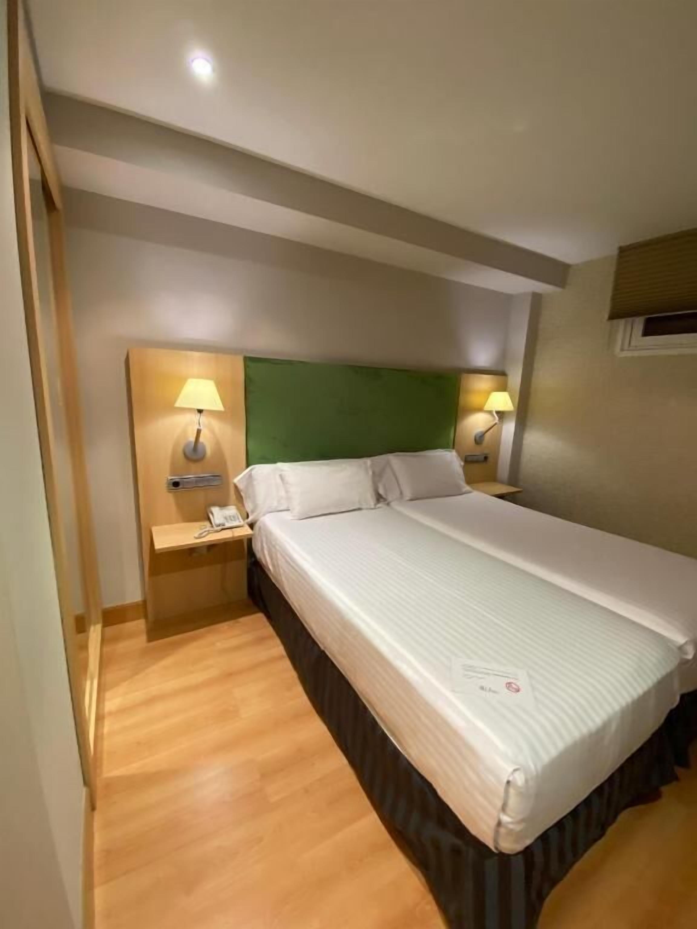 Habitación de hotel con una cama grande, dos mesitas de noche con lámparas, cabecera verde y suelo de madera.