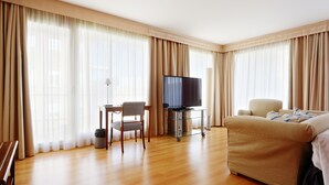 Habitación de hotel con piso de madera, un sofá, una silla, un escritorio, un televisor y ventanas grandes con cortinas.