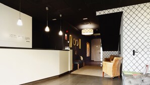 Un área de recepción con un cartel que dice "HOTEL VILLA GOMA", un mostrador de recepción, luces colgantes, un muro con diseño y un espacio de descanso con sofá y sillones.