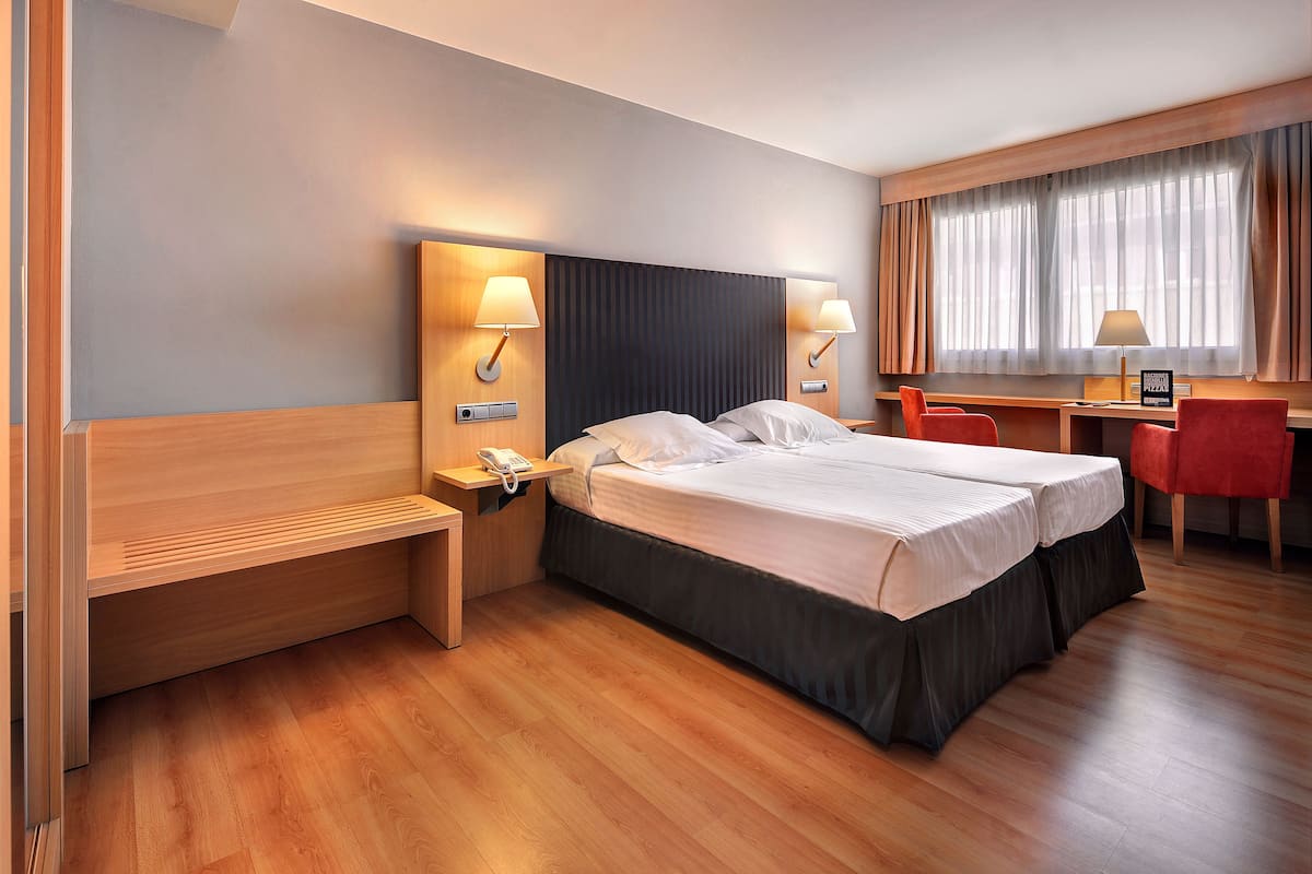 Habitación de hotel con cama doble, escritorio con silla, teléfono y ventana con cortinas.