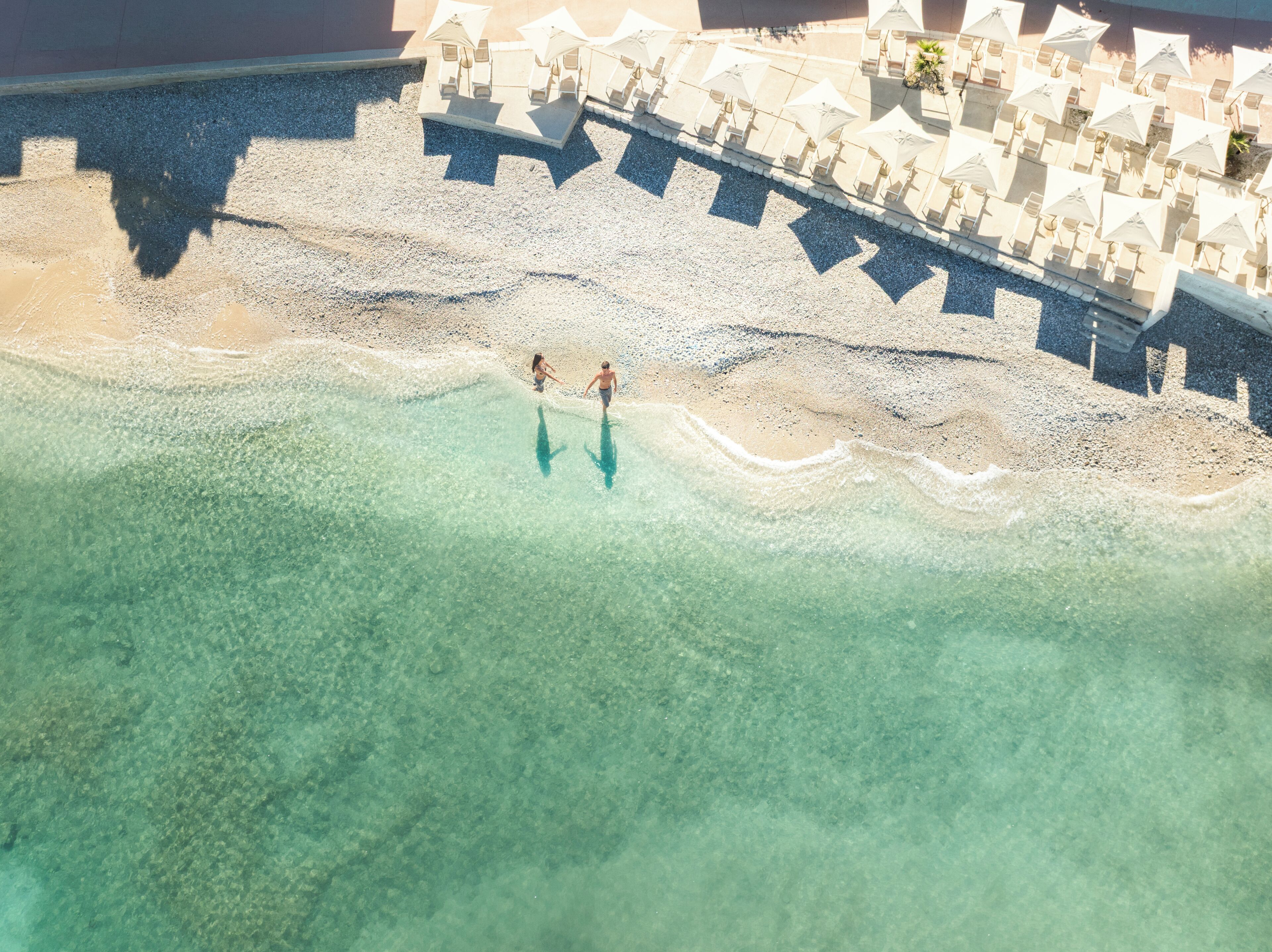 Vista despejada de una playa con dos personas caminando en el agua, una hilera de sombrillas y un edificio al fondo.