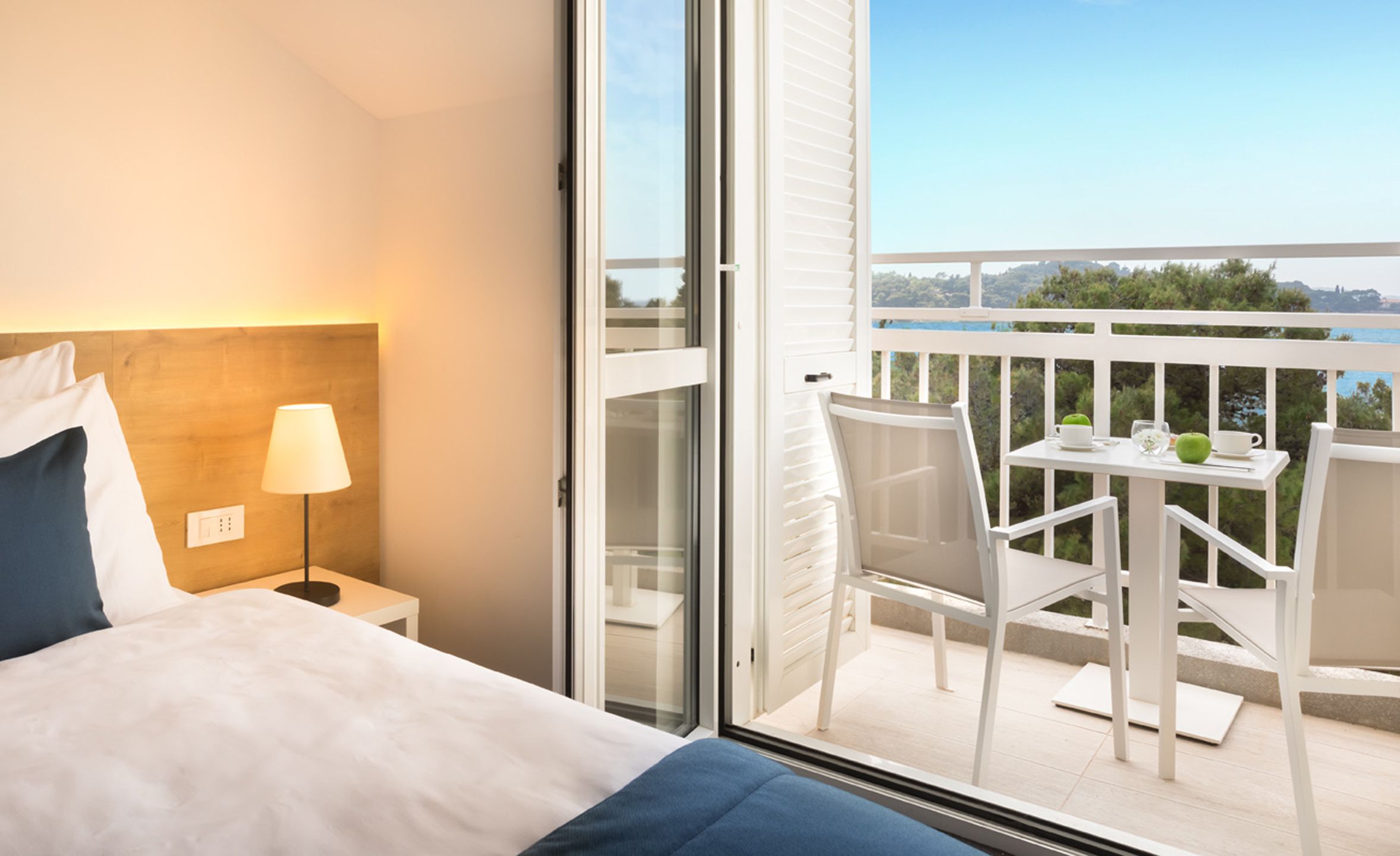 A hotel room with a bed, a nightstand with a lamp, and a balcony with a table and chairs.