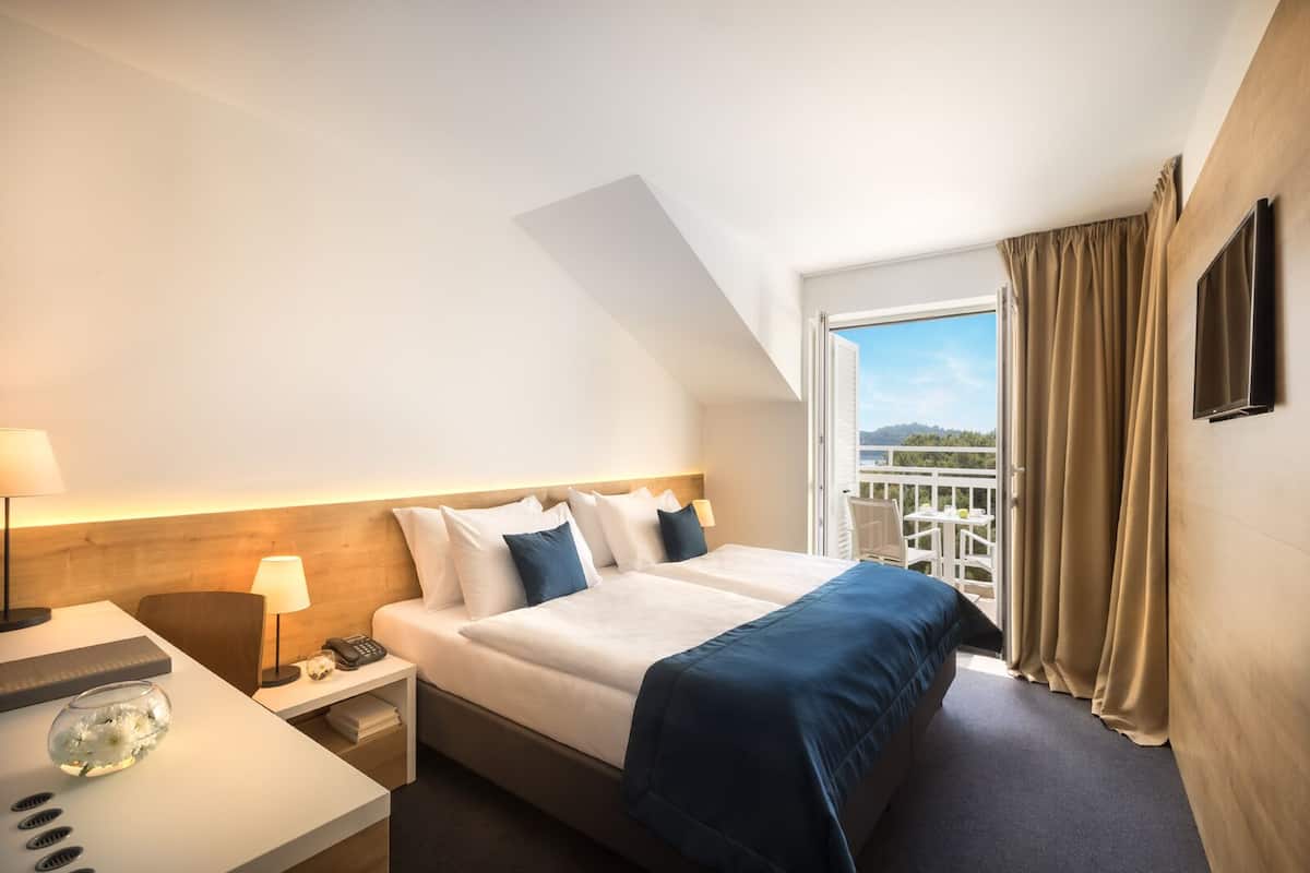 A hotel room with a bed, bedside table, lamp, and a view of the outdoors.