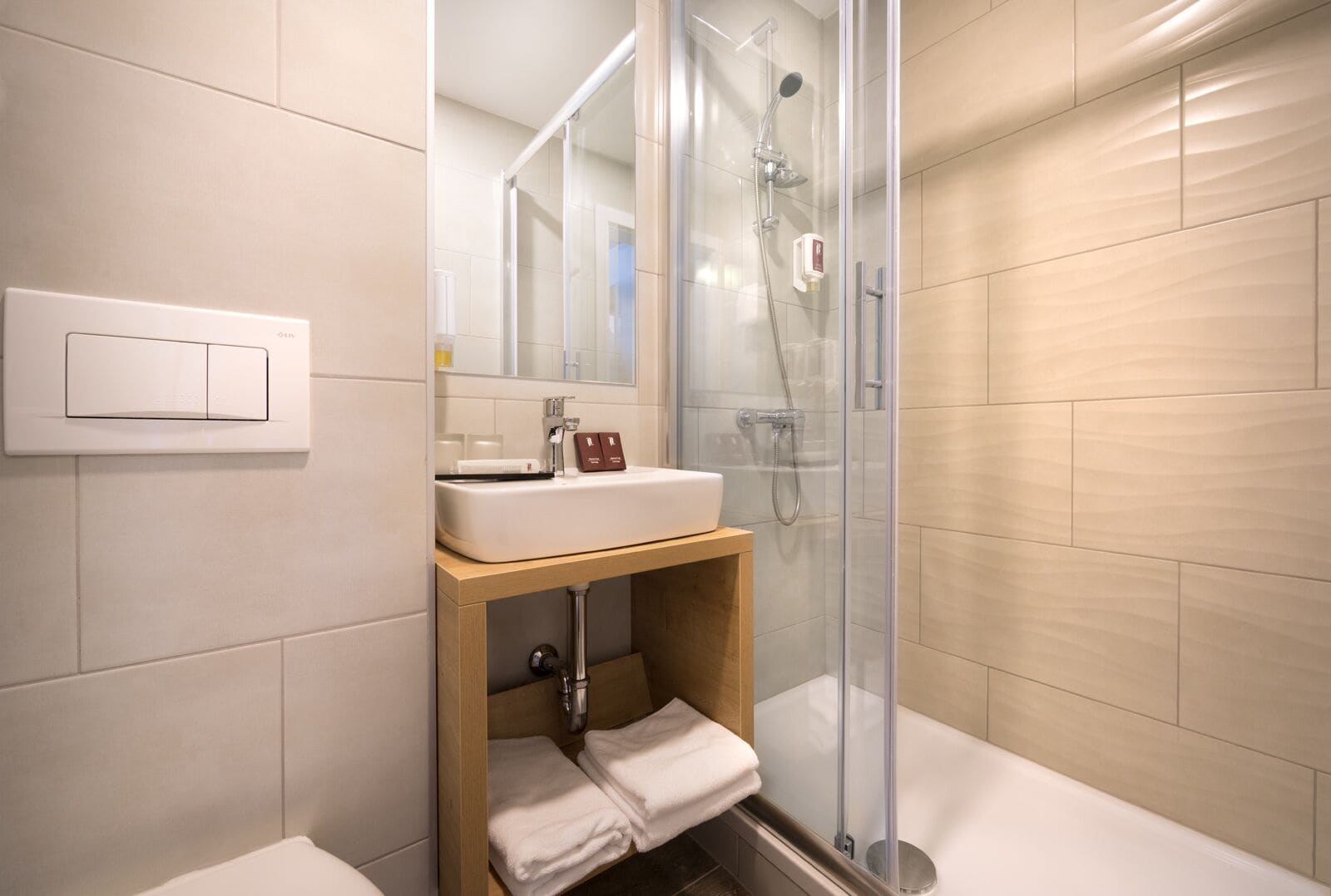 Un baño moderno con ducha envuelta en vidrio, un lavabo blanco con espejo y un estante de madera con toallas.