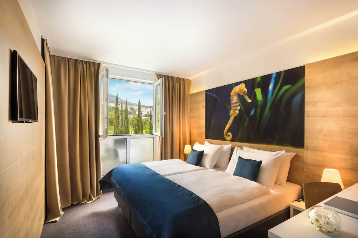 A hotel room with a large bed, a TV, a window with curtains, a painting, and a nightstand with a lamp.