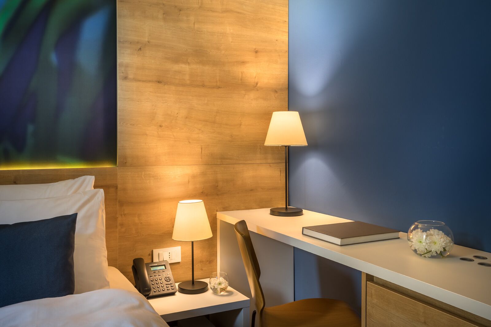 A hotel room with a bed, bedside table with a lamp, a desk with a chair, a phone, and a vase with flowers.