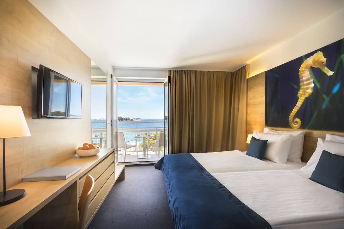 A hotel room with a large bed, a desk with a lamp, a chair, and a view of the sea through the window.