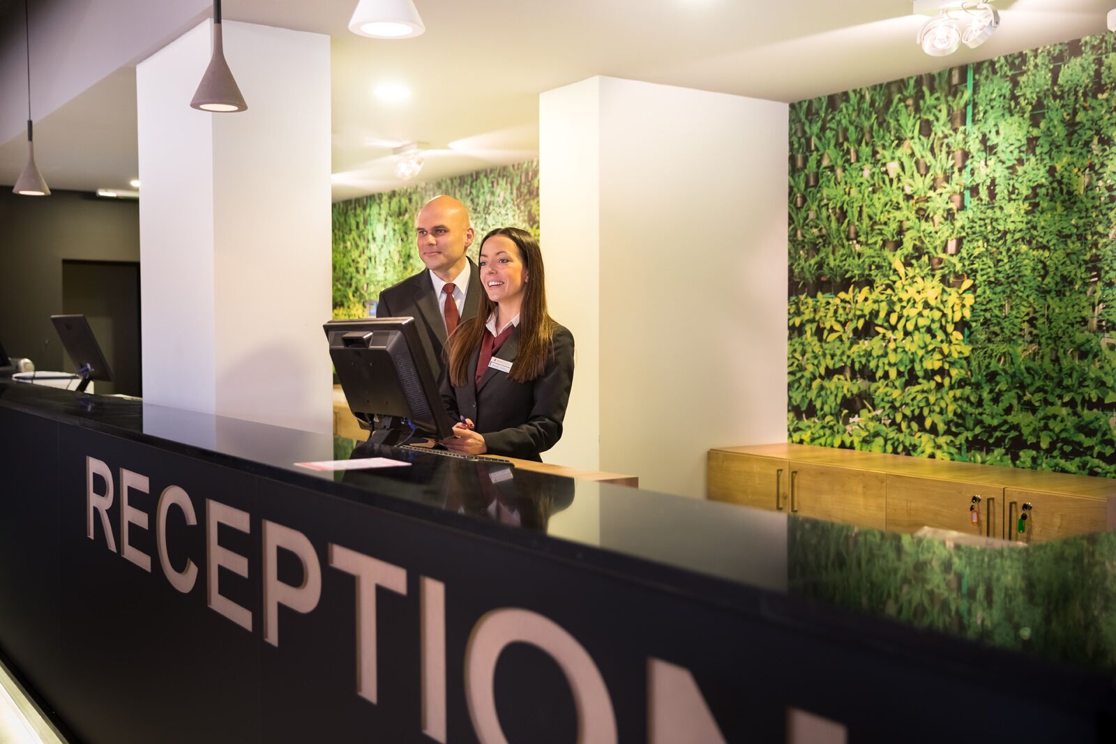 Ein Empfangstisch mit einer Namensschild, auf der „RECEPTION“ steht, und eine Wand mit einem grünen, blattartigen Muster im Hintergrund.