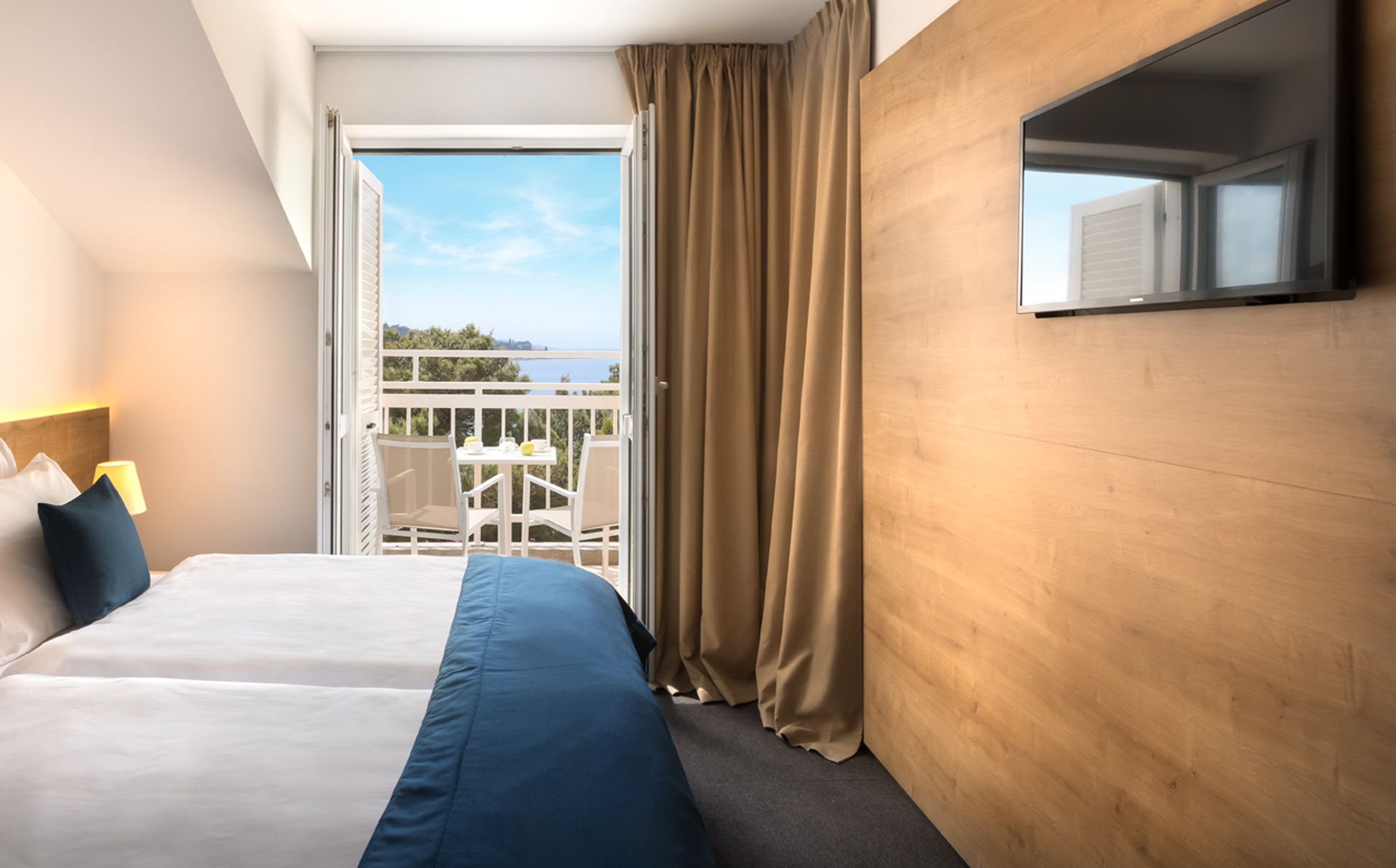 A hotel room with a bed, a TV mounted on the wall, and a balcony with a view of the sea.