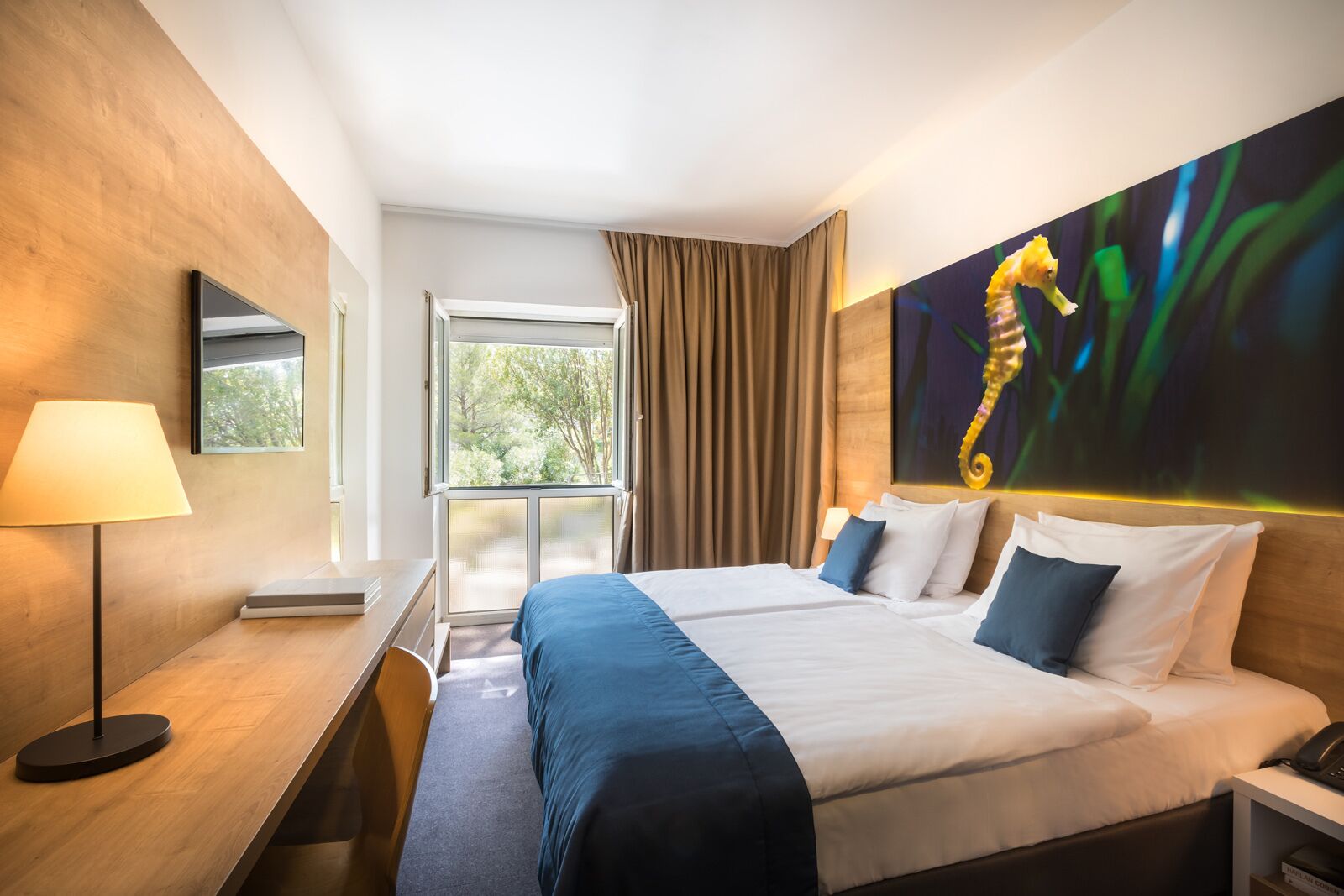 Hotelkamer met een groot bed, een bureau, een lamp, een raam met gordijnen en een schilderij van een zeepaard aan de muur.