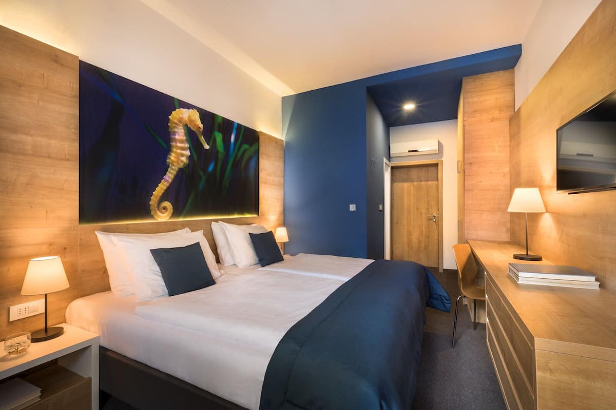 A modern hotel room with a large bed, a desk, a TV, and a seahorse painting on the wall.