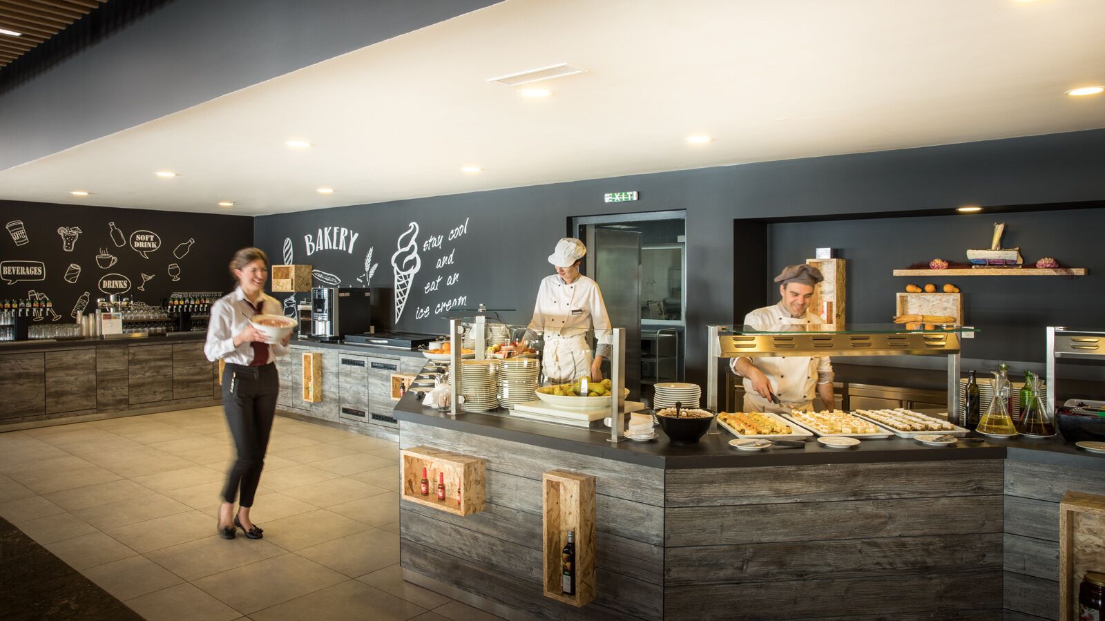 Mostrador de panadería con chefs preparando alimentos, zona de bar con bebidas y aperitivos, y una pared con ilustraciones y texto relacionados con la comida.