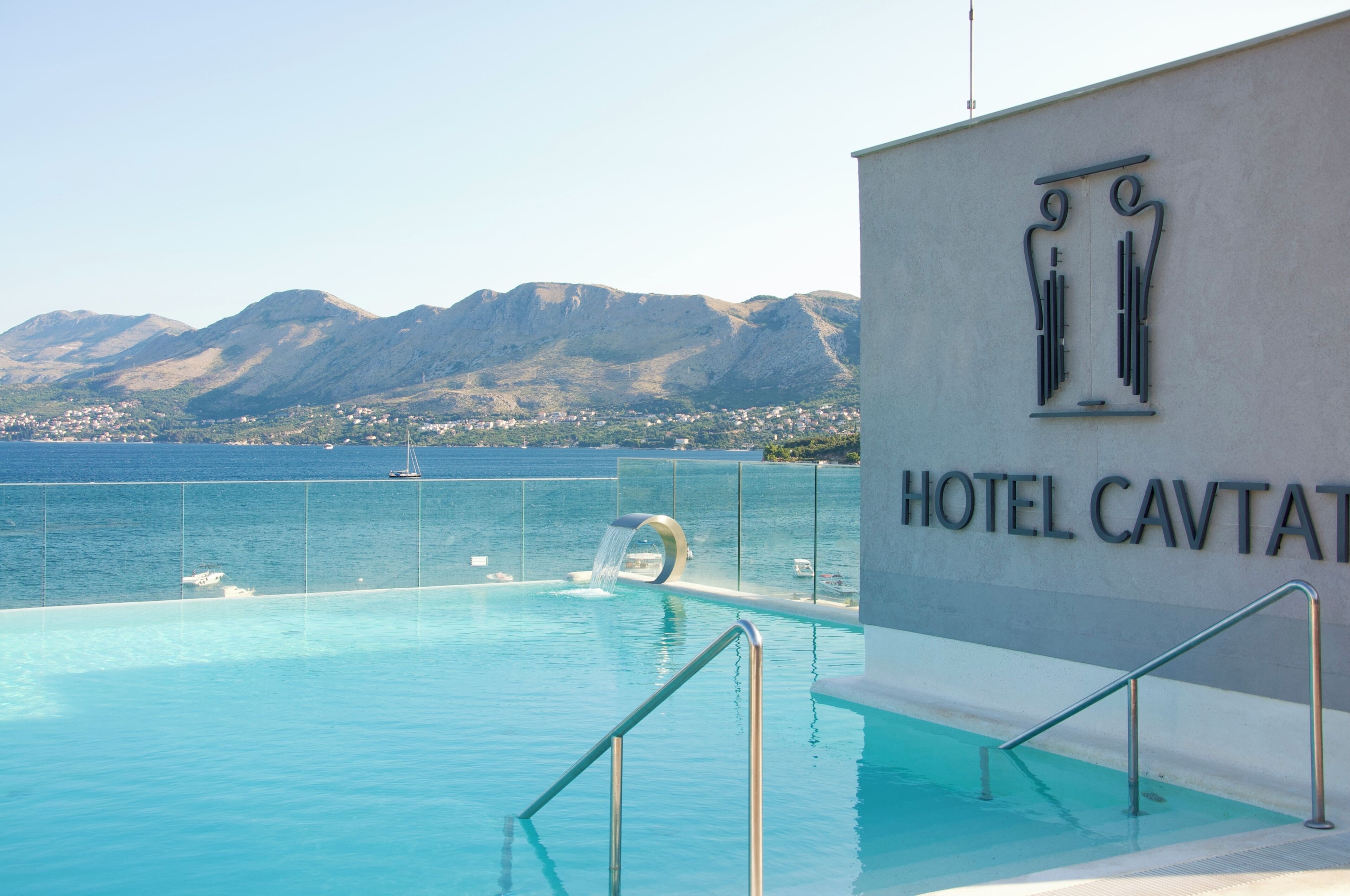 Una piscina con vistas al mar y las montañas, un cartel de Hotel Cavtat, y un diseño arquitectónico moderno.