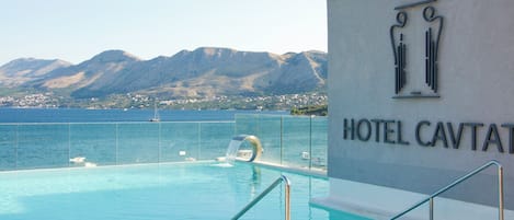 A swimming pool with a view of the sea and mountains, a sign for Hotel Cavtat, and a modern architectural design.