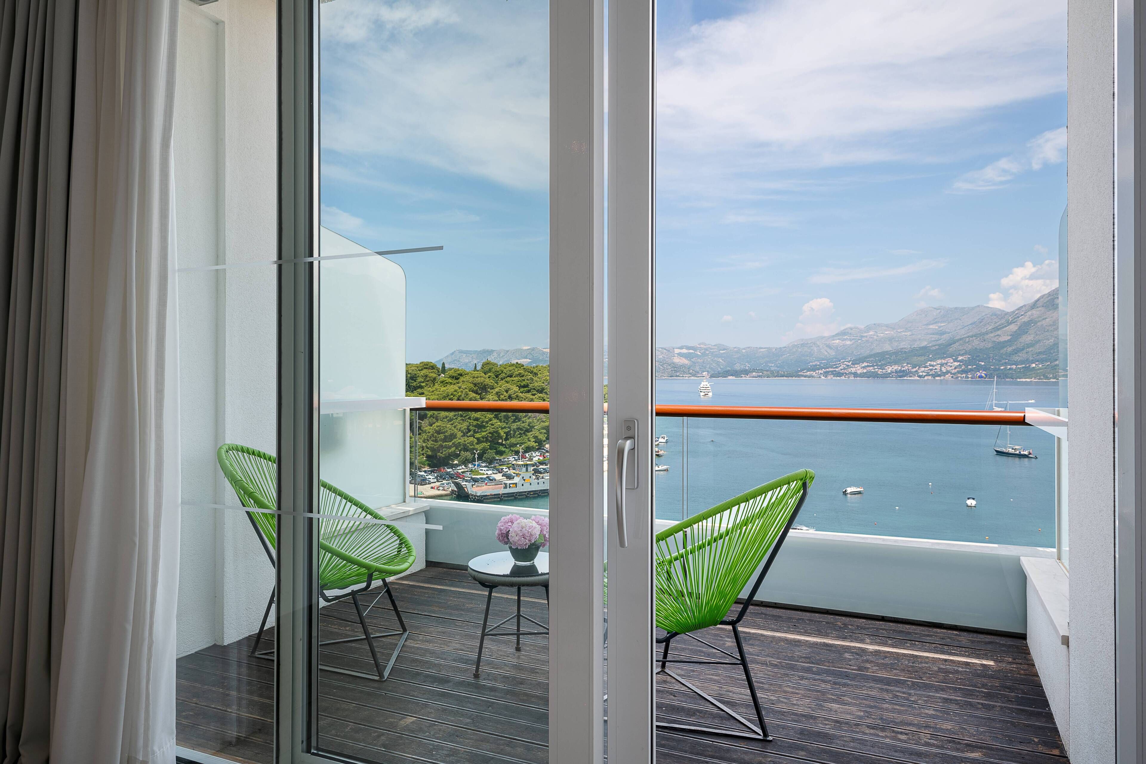 A balcony with a view of the sea, two green chairs, and a small table with flowers.