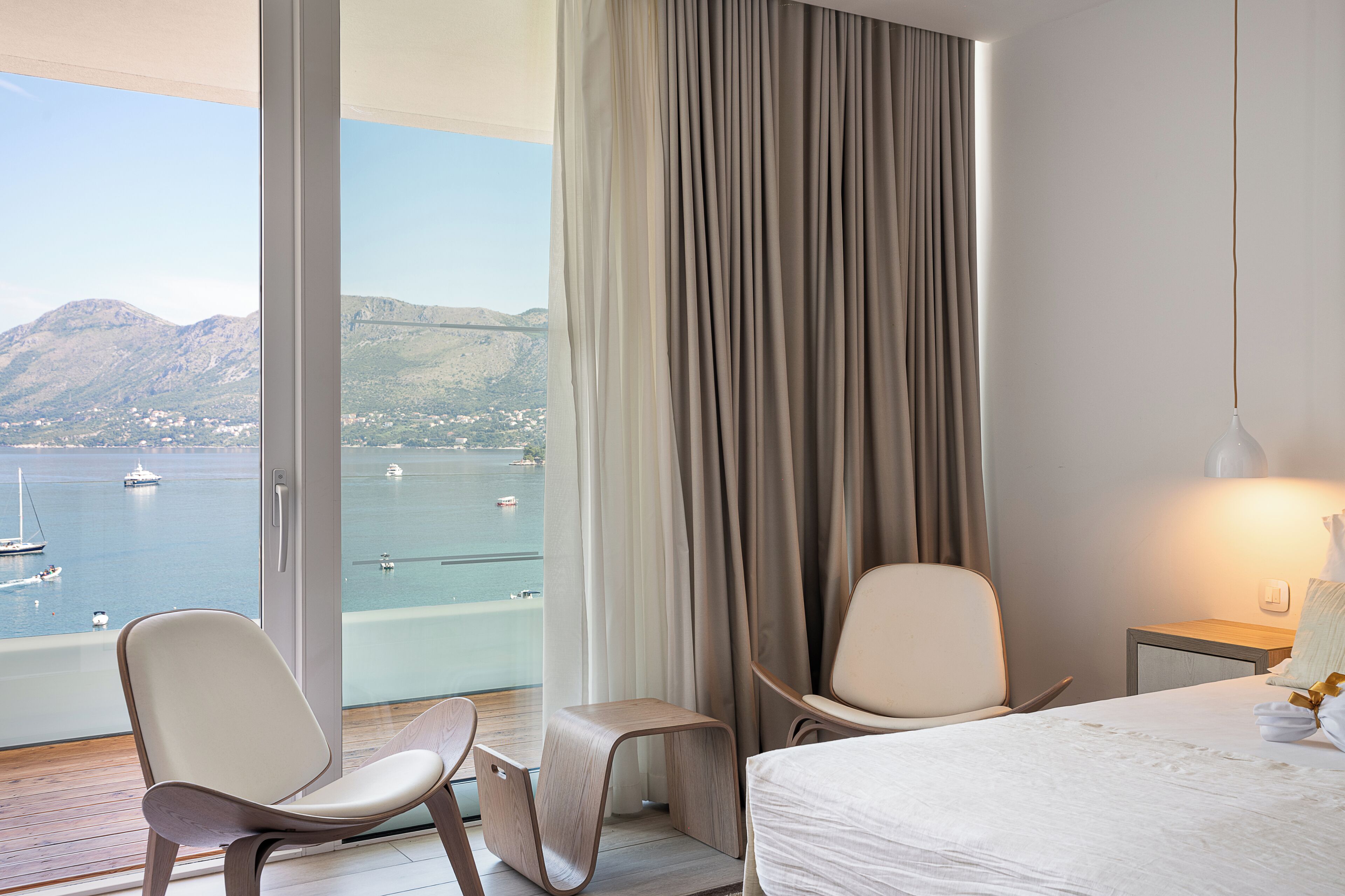 A hotel room with a large window offering a view of a body of water and mountains. There is a bed, two chairs, and a small table.