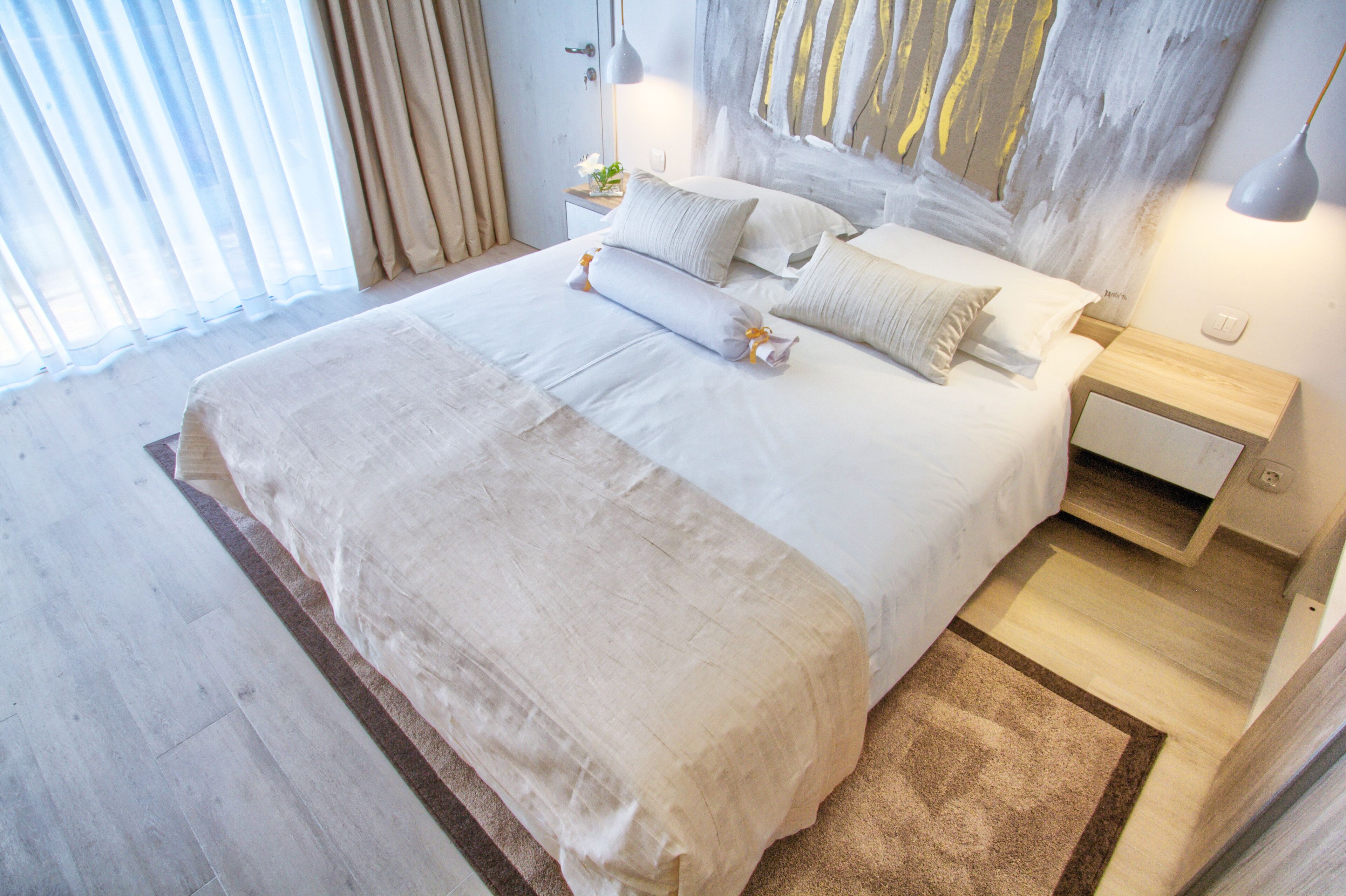 Uma cama bem arrumada com um edredom branco grande e travesseiros, uma mesa de cabeceira de madeira com uma luminária e um quadro abstrato moderno na parede.