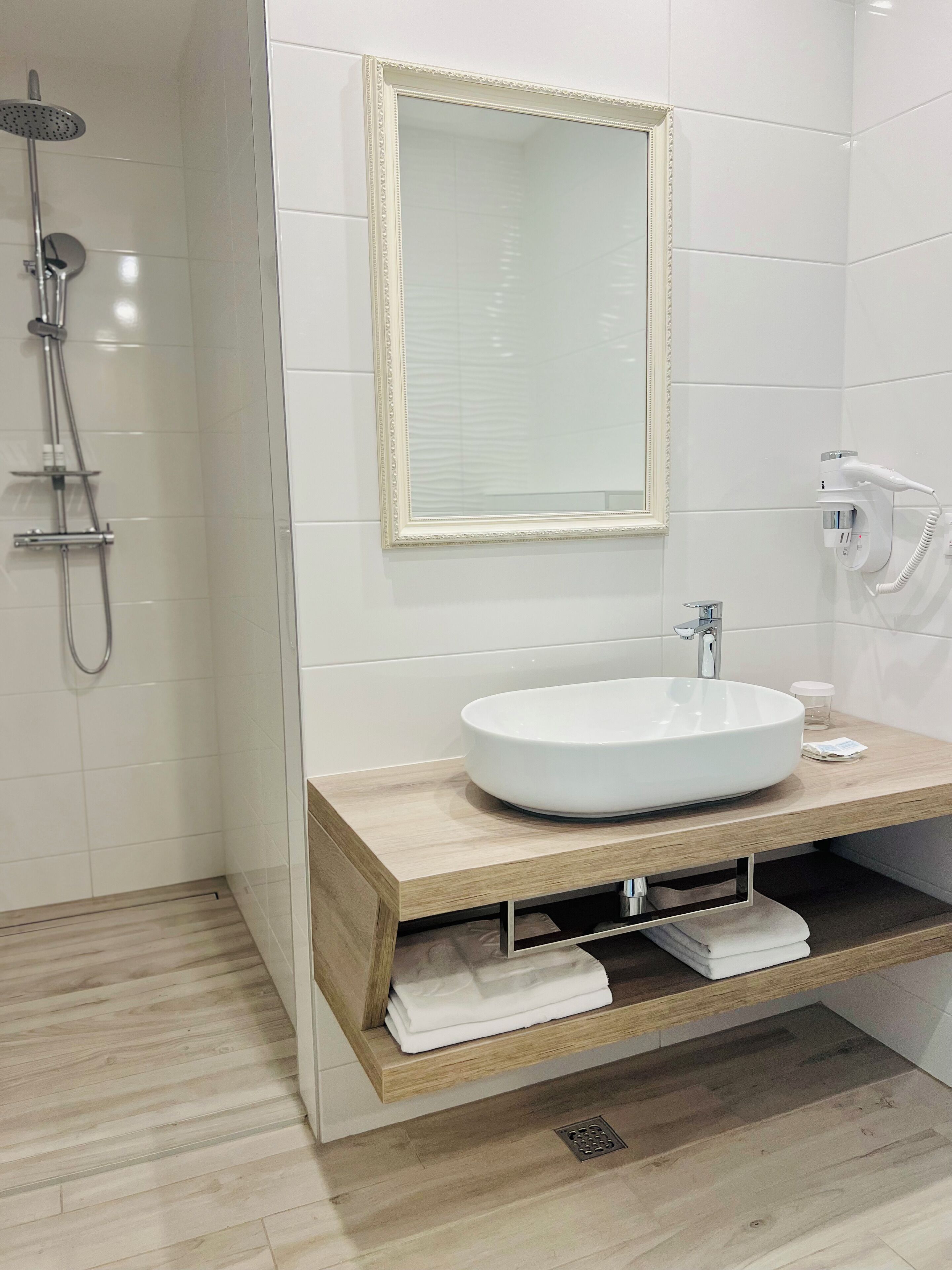 A modern bathroom with a large white sink, a wooden vanity, a mirror with a decorative frame, a handheld showerhead, and a hairdryer mounted on the wall.