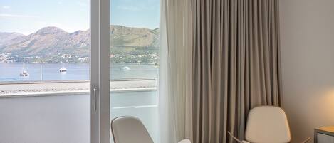 Ein Hotelzimmer mit großem Fenster, das auf ein Gewässer und Berge Blick bietet, einem Bett, zwei Stühlen und einem Vorhang.