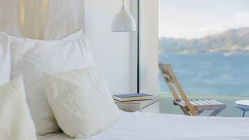 A bedroom with a bed, pillows, a bedside table with a book, a lamp, and a view of the sea.