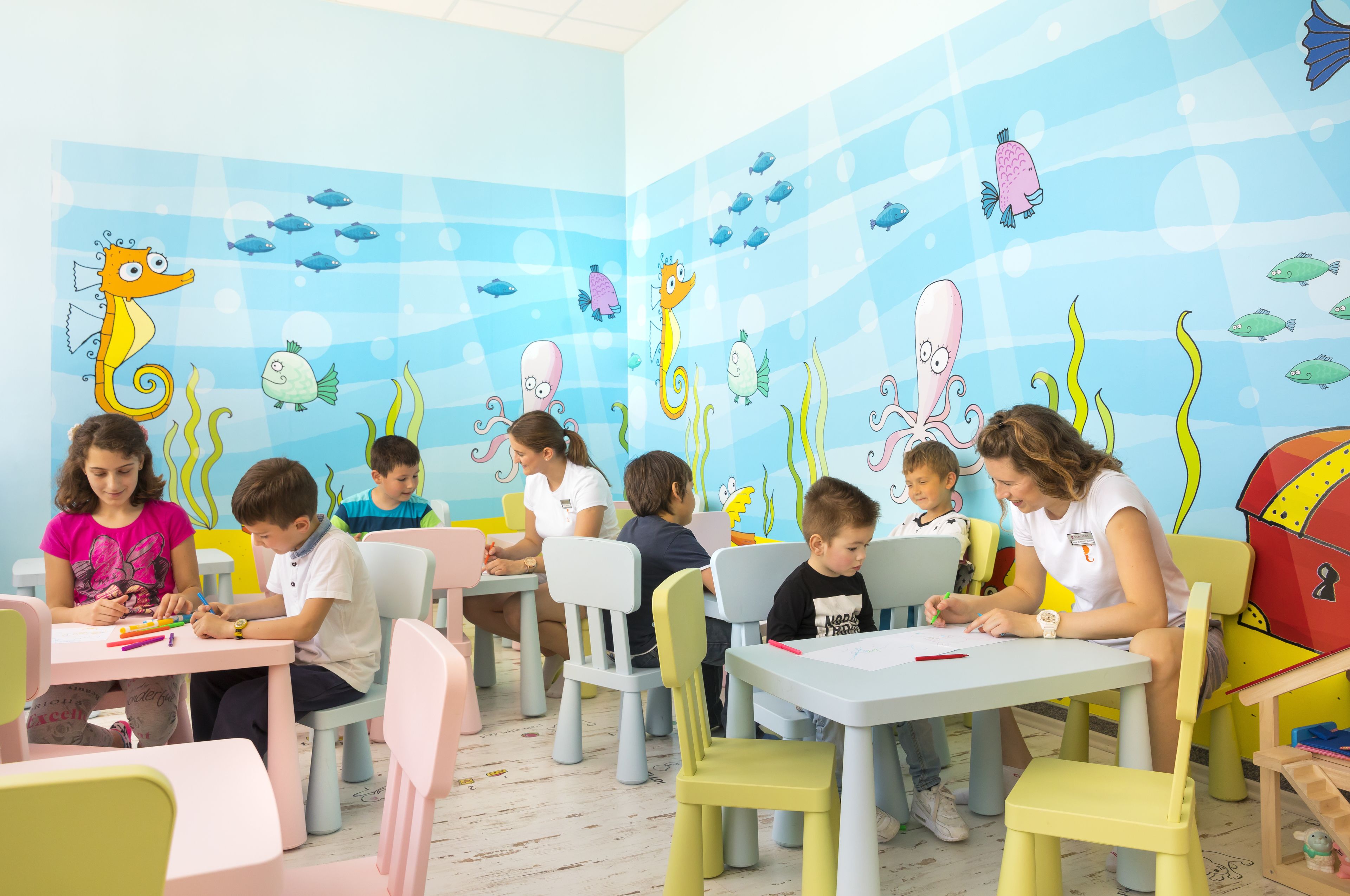 Une salle de classe où des enfants sont assis à des tables, occupés à diverses activités, et où les murs sont décorés d’une fresque colorée représentant un paysage sous-marin.