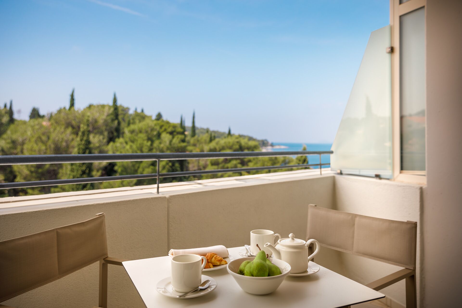 A balcony with a table set for breakfast, including a bowl of pears, a croissant, and a teapot.
