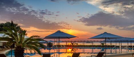 A poolside area with lounge chairs and umbrellas, overlooking a body of water with a sunset in the background.