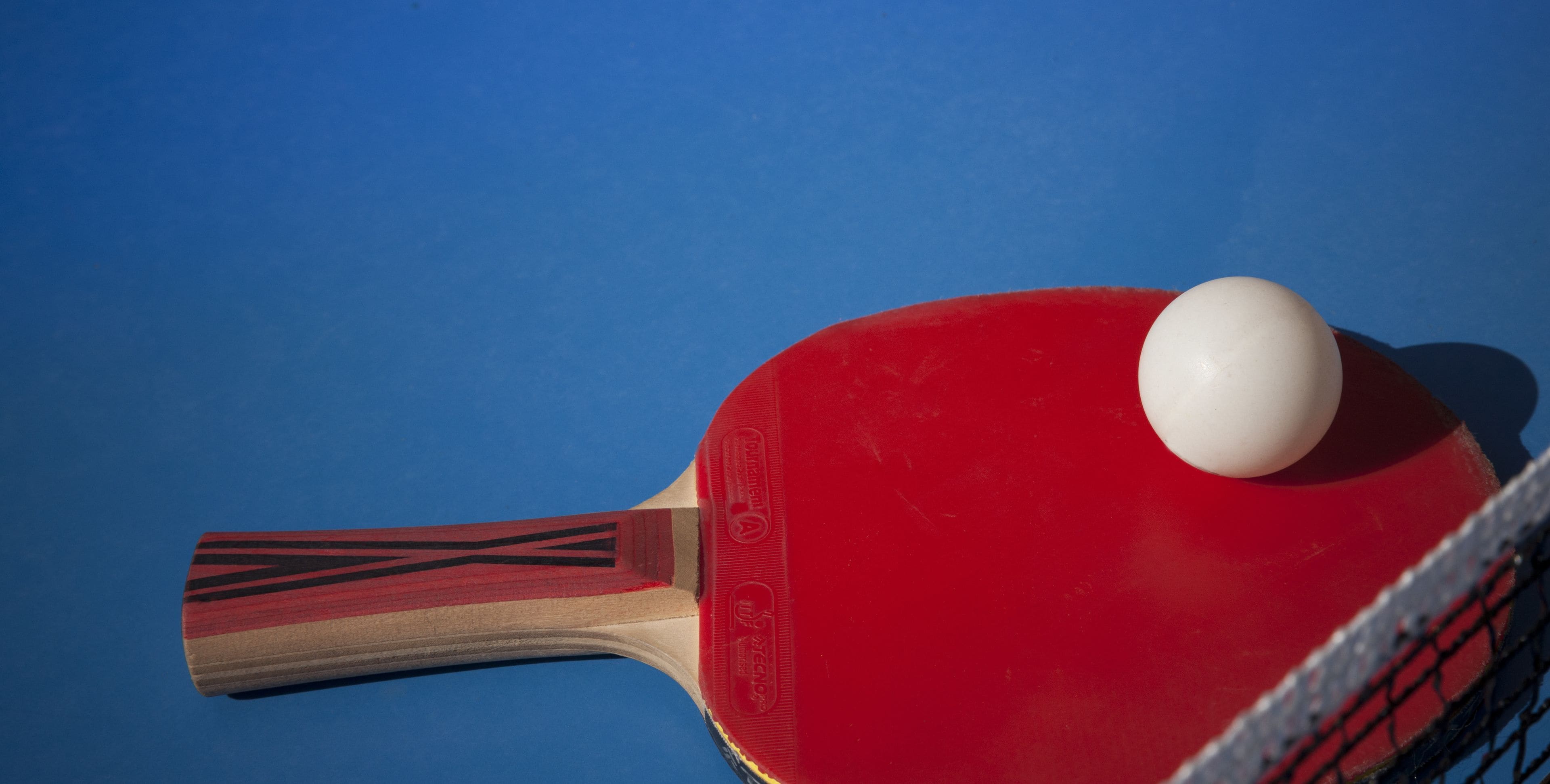 Una paleta de ping pong con una pelota roja y blanca sobre una mesa azul.