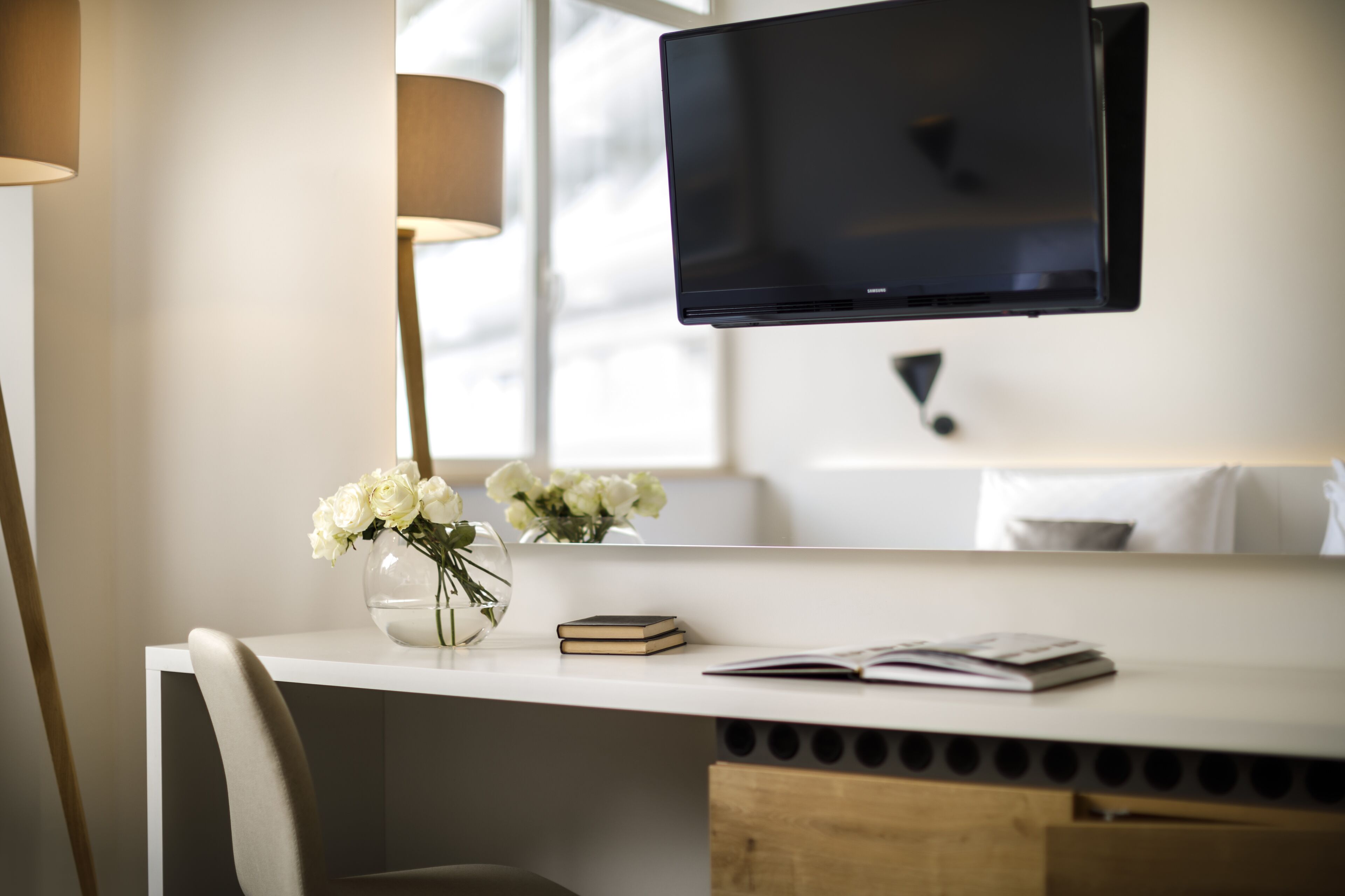 Una habitación moderna con un escritorio blanco, un televisor de pantalla plana en la pared, un florero con flores blancas y una lámpara.