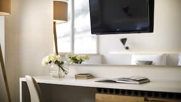 Una habitación moderna con un escritorio blanco, un televisor de pantalla plana en la pared, un florero con flores blancas y una lámpara.
