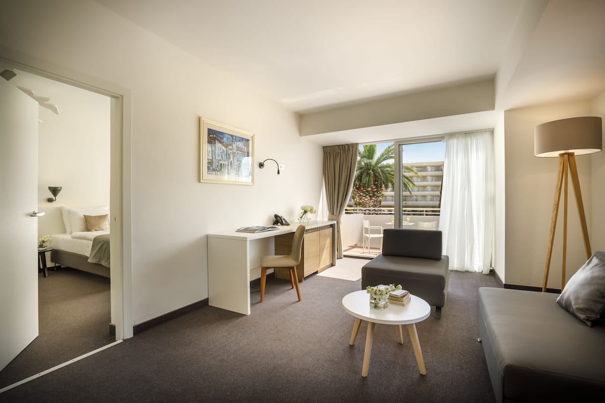 Ein modernes Hotelzimmer mit Schreibtisch, Sofa, einem kleinen Couchtisch und einem Balkon mit Blick auf Palmen.