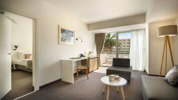 Habitación de hotel moderna con escritorio, sofá, una pequeña mesa de centro y balcón con vista a palmeras.