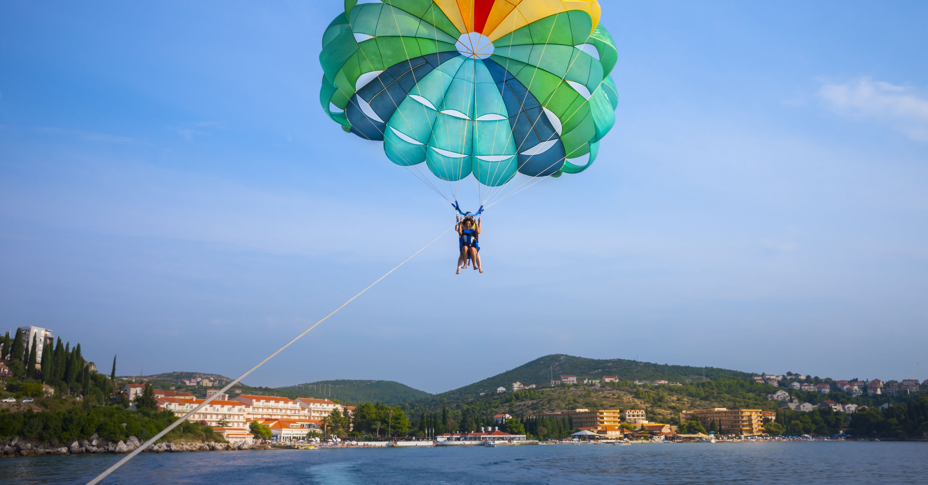 Une personne pratiquant le parasailing au-dessus de l’eau, avec des bâtiments et des collines en arrière-plan.