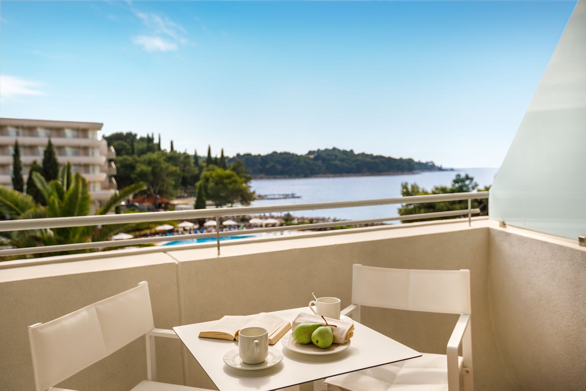 Un balcón con una mesa para dos, vista a un cuerpo de agua y una costa lejana con edificios y árboles.