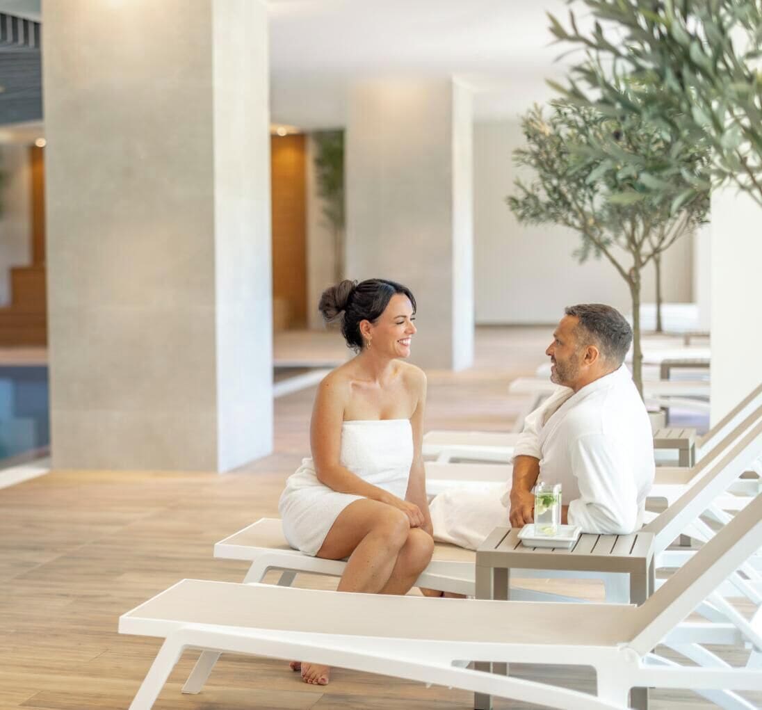 Una pareja sentada en sillones de sol en un espacio interior moderno, con suelo de madera y un árbol grande al fondo.
