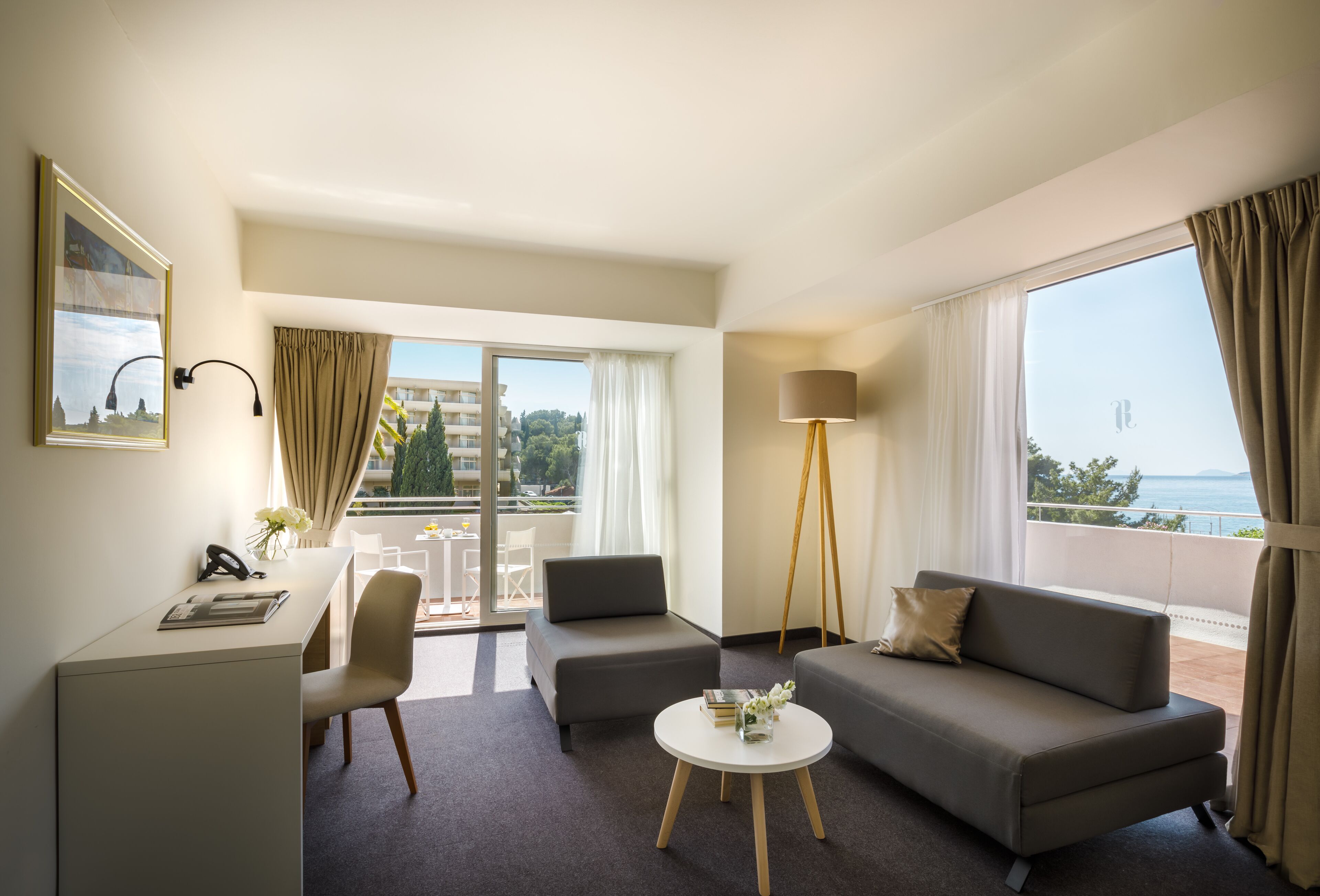 Quarto de hotel moderno com varanda, uma escrivaninha, dois sofás e um abajur.