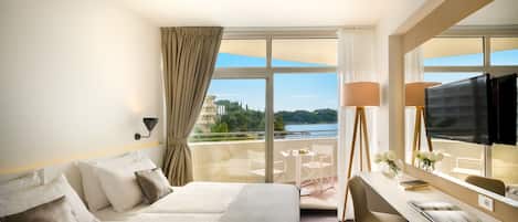 A hotel room with a bed, a desk, a TV, and a balcony with a view of the water.