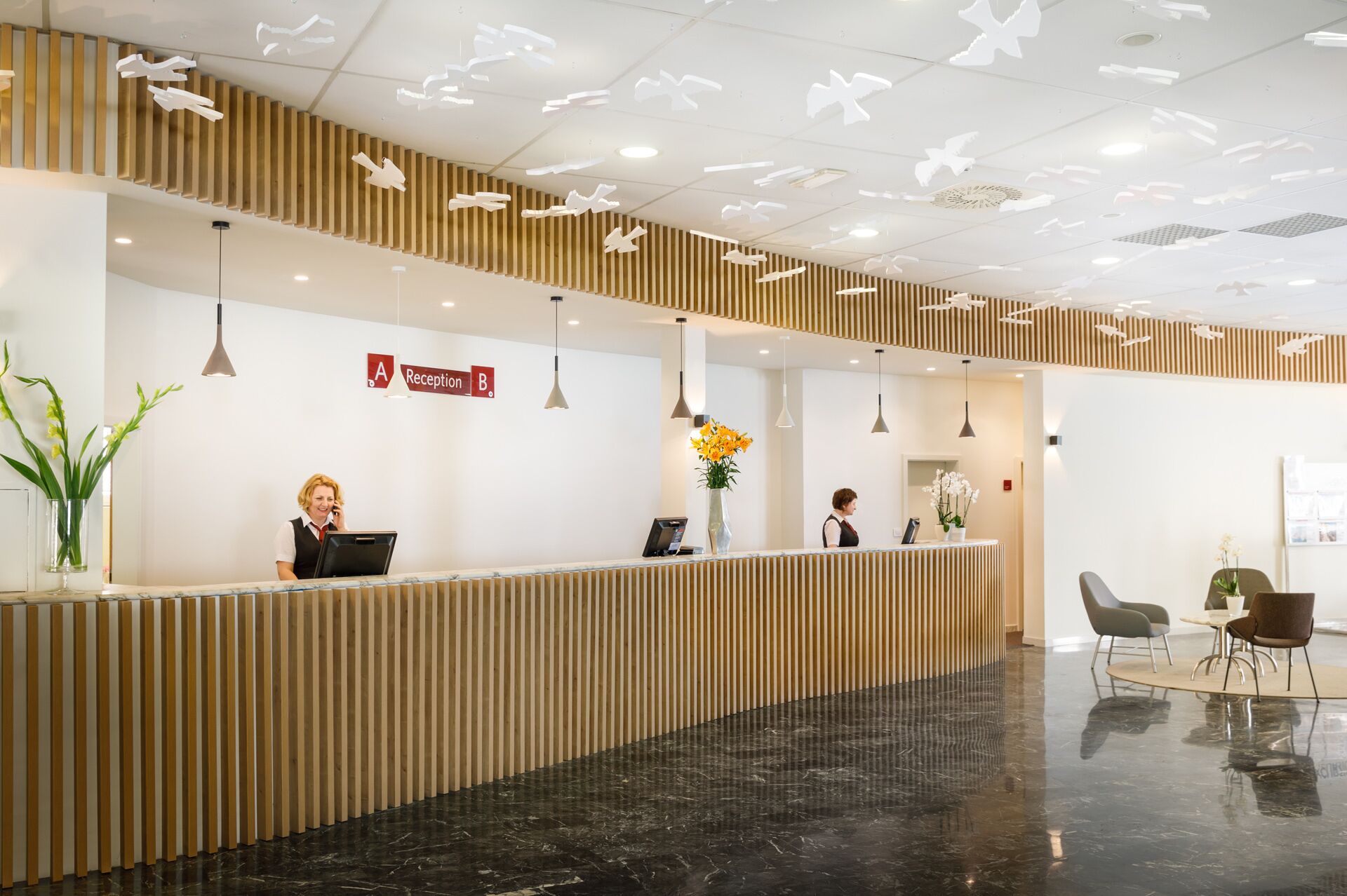 A reception area with a wooden reception desk, a sign indicating reception areas A and B, and a modern ceiling design with hanging light fixtures.