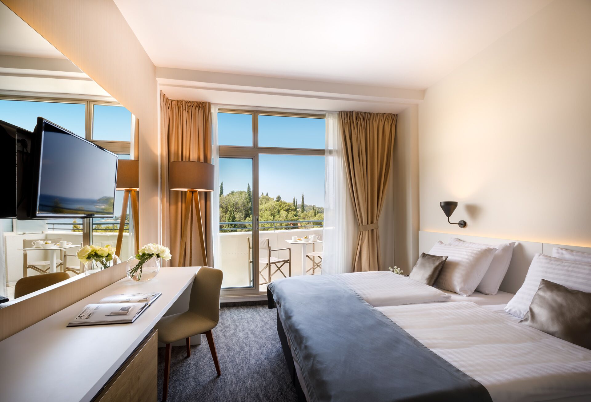 Habitación de hotel con cama, escritorio con televisor, balcón con mesa y sillas, y vista a zonas verdes.