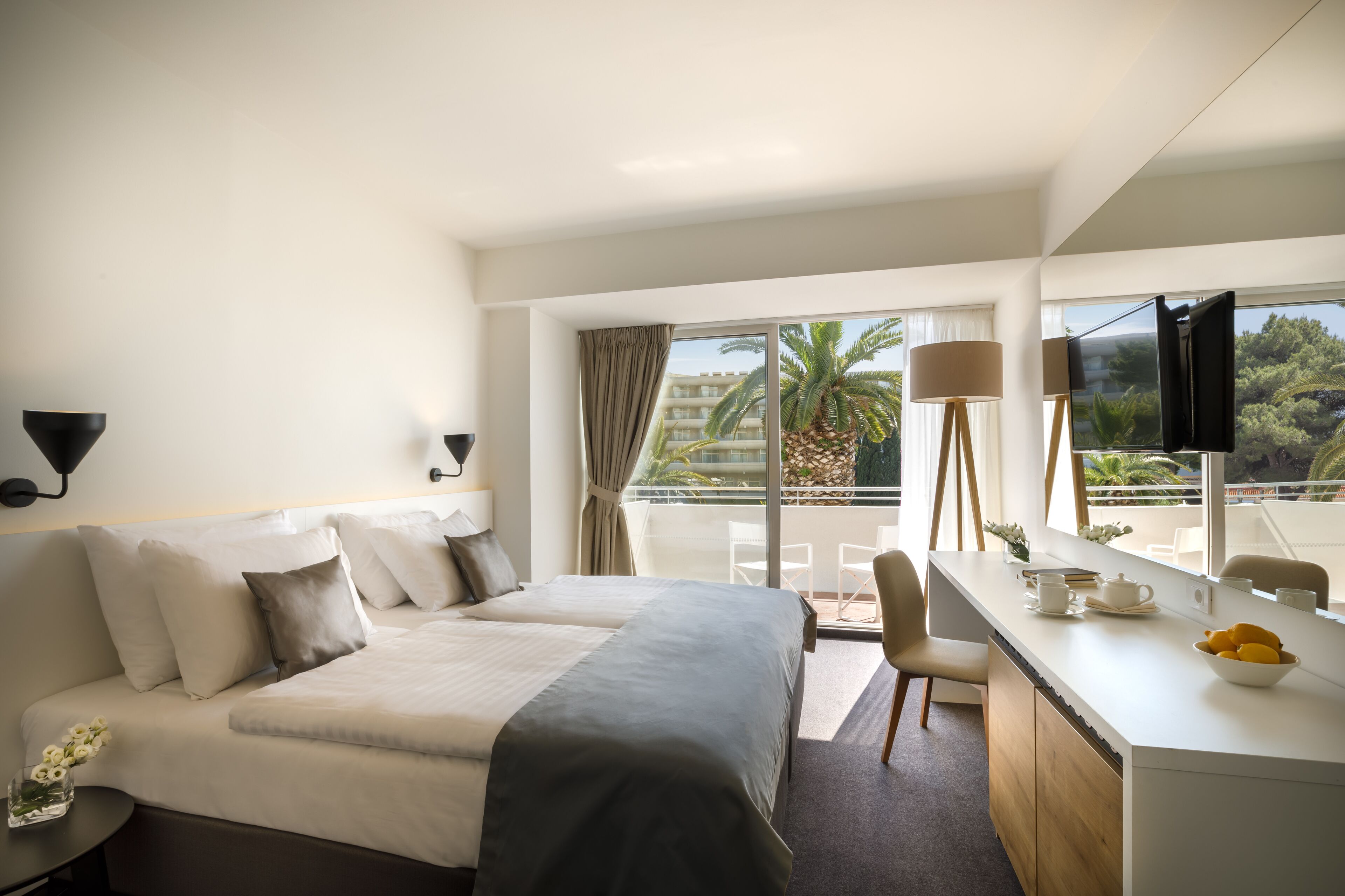 Habitación de hotel con una cama grande, un escritorio con un frutero, un televisor y un balcón con vista a palmeras y un edificio.